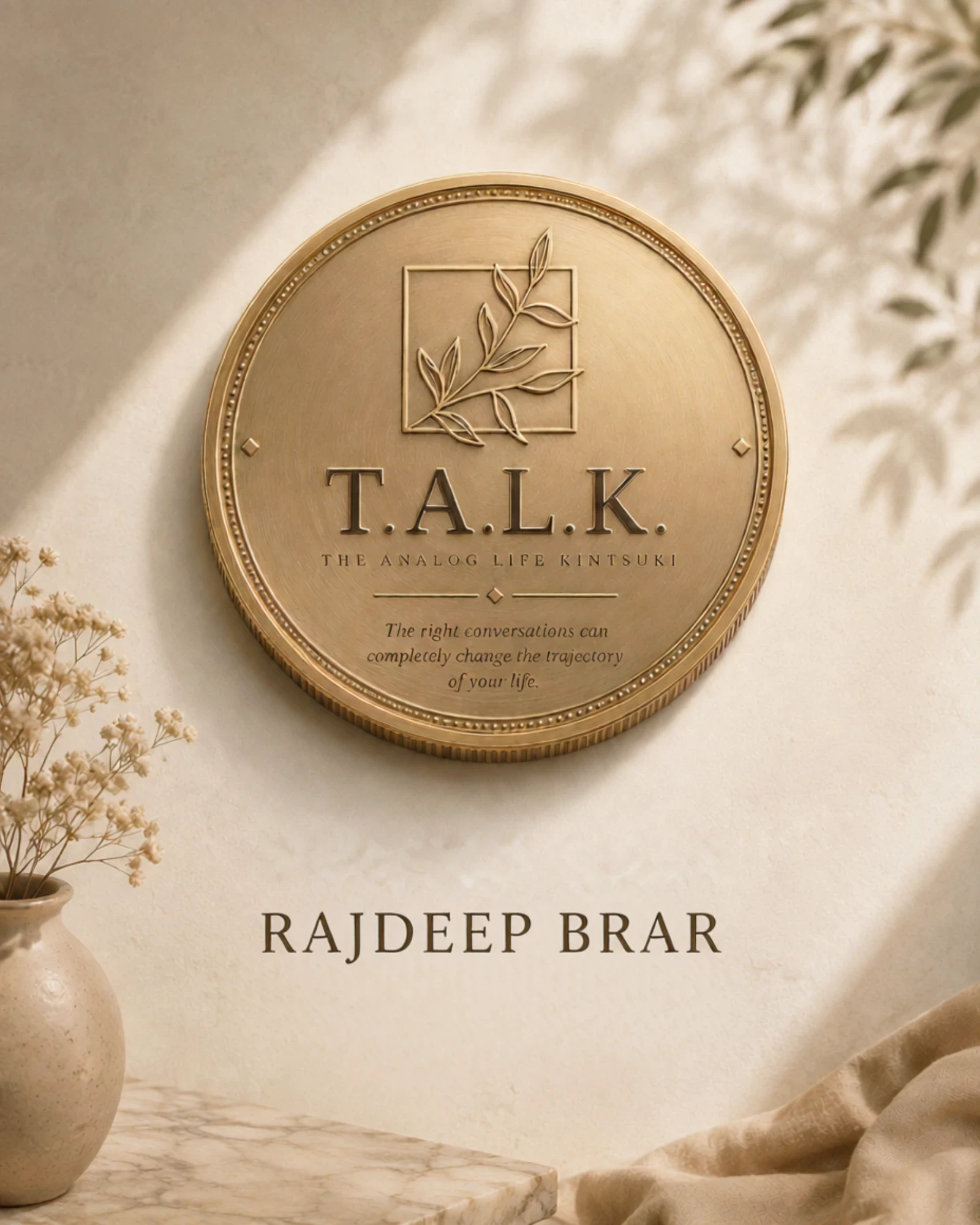 A cast brass T.A.L.K. medallion on a warm plaster wall beside dried flowers
